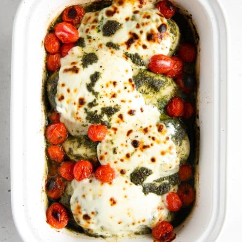 White casserole dish filled with four large oven-baked chicken breasts smothered in pesto sauce, fresh melted mozzarella cheese, and burst cherry tomatoes.