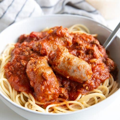 Image of a white bowl filled with spaghetti covered with spaghetti sauce made with ground beef and topped with sausage.