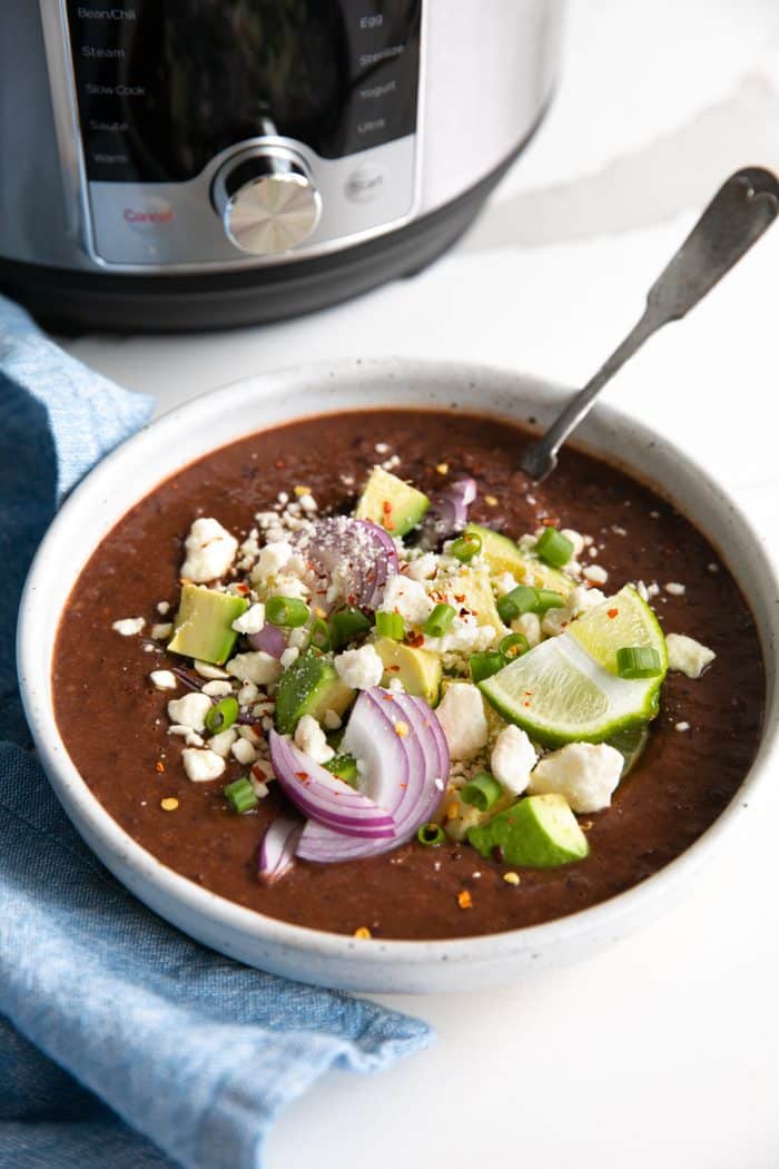 Large shallow soup bowl filled with Instant Pot black bean soup and topped with crumbled white cheese, avocado, red onion, red chili flakes, and fresh lime wedge.