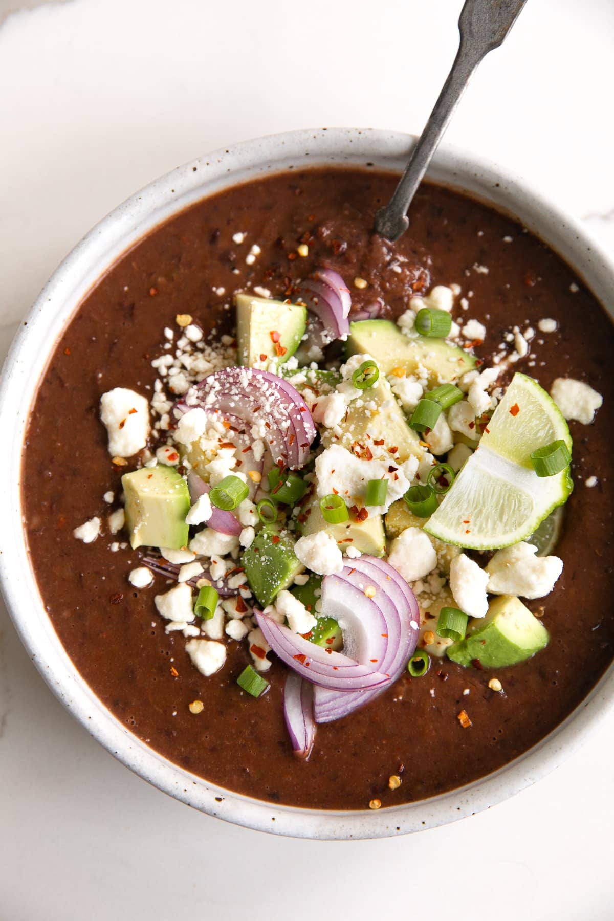 Overhead image of a large shallow soup bowl filled with Instant Pot black bean soup and topped with crumbled white cheese, avocado, red onion, red chili flakes, and fresh lime wedge.