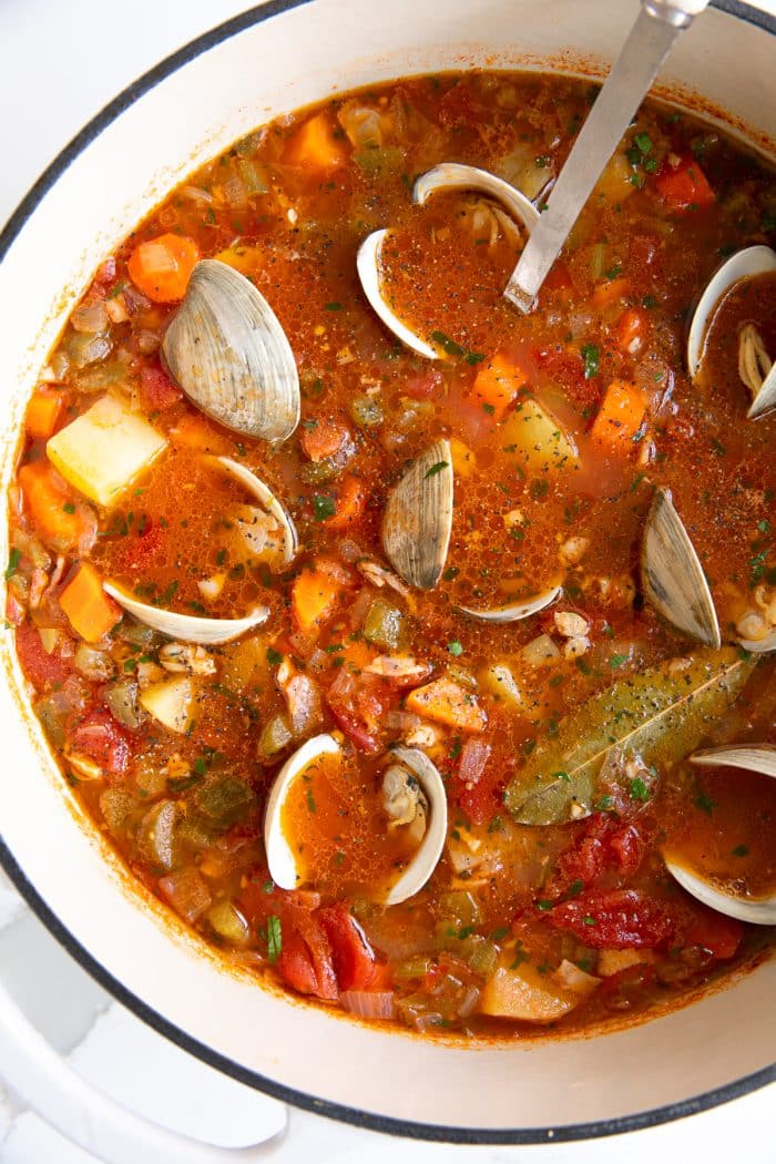 Overhead image of a large pot of Manhattan clam chowder filled with carrots, celery, onion, potatoes, little neck clams, all simmering in a tomato and clam broth.