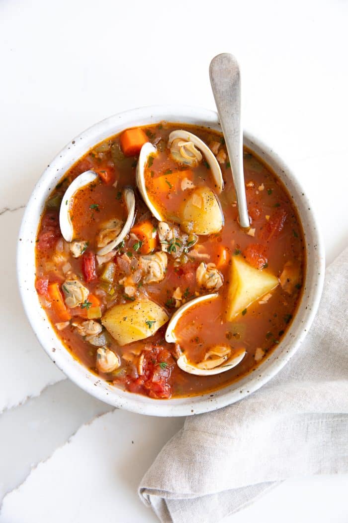 Overhead image of a white soup bowl filled with homemade Manhattan clam chowder recipe made with little neck clams, potatoes, carrots, in a light tomato clam broth.