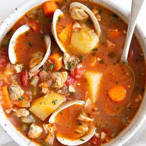 Overhead image of a white soup bowl filled with homemade Manhattan clam chowder recipe made with little neck clams, potatoes, carrots, in a light tomato clam broth.