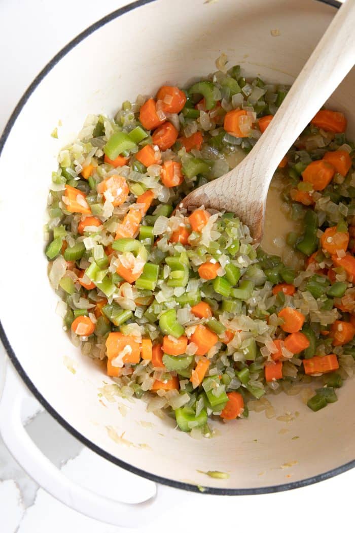 Diced onion, carrots, and celery cooking in a large white ceramic pot.