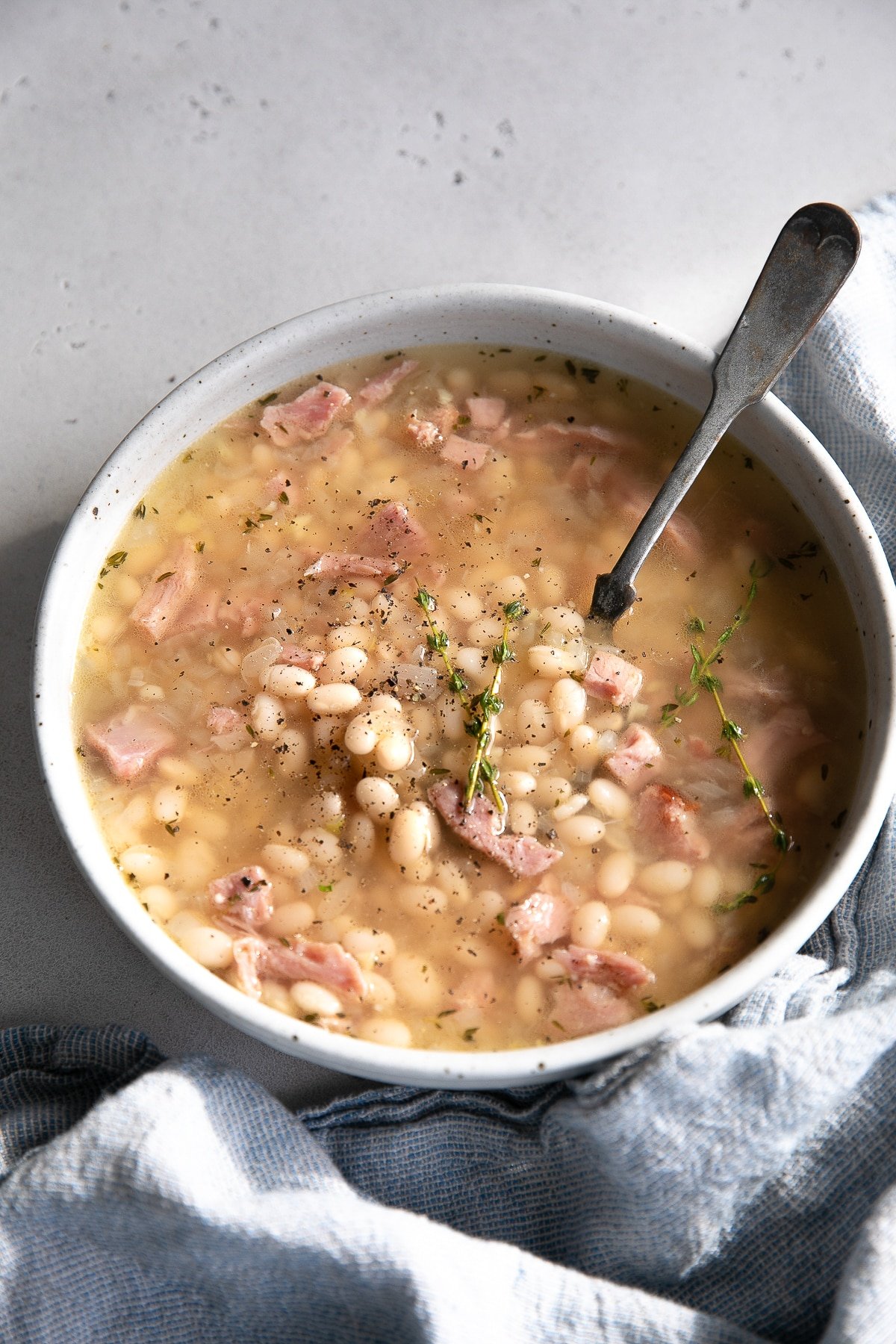 White soup bowl filled with navy bean soup made with chunks of leftover ham, cooked navy beans, and fresh thyme in a light broth.