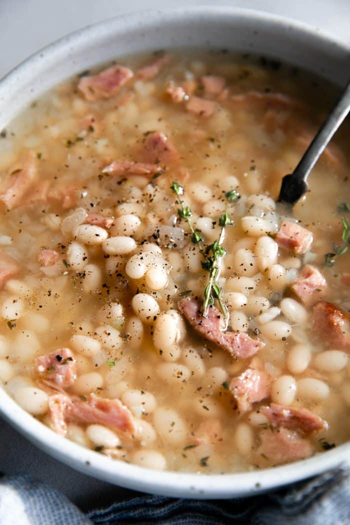 White soup bowl filled with navy bean soup made with chunks of leftover ham, cooked navy beans, and fresh thyme in a light broth.