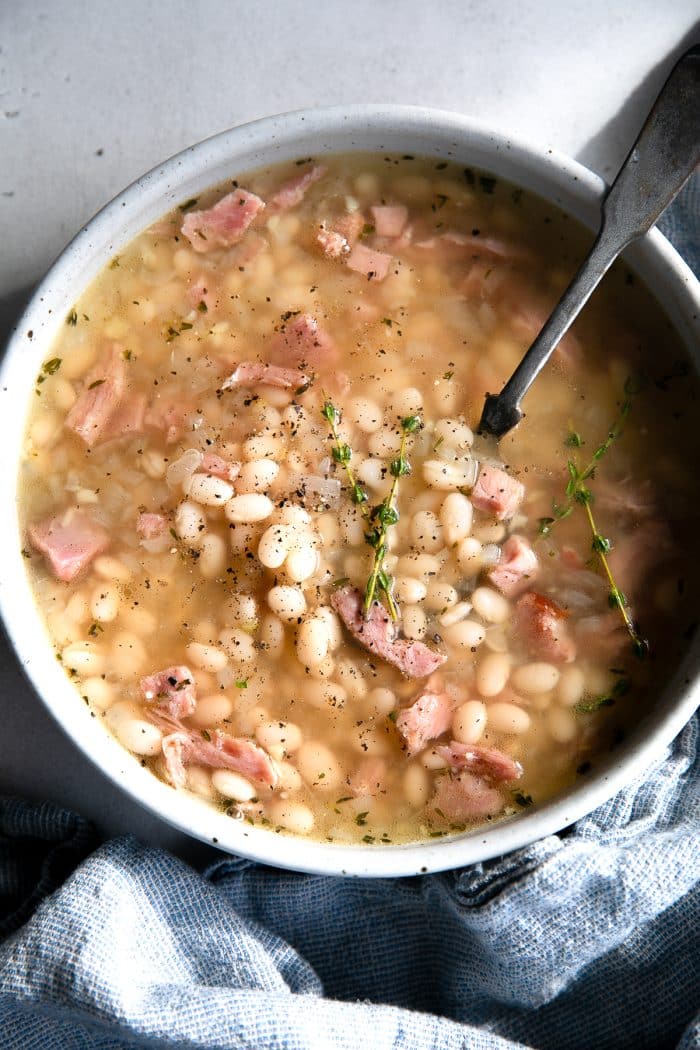 White soup bowl filled with navy bean soup made with chunks of leftover ham, cooked navy beans, and fresh thyme in a light broth.