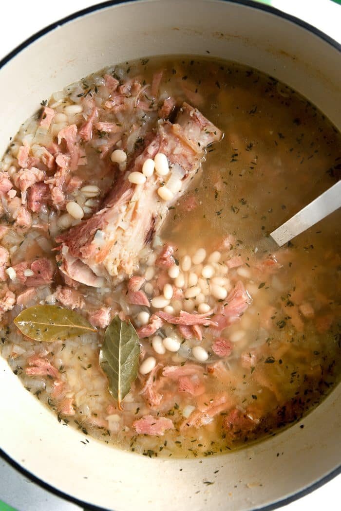 Large soup pot filled with navy beans, leftover ham, and ham bone in a light broth.