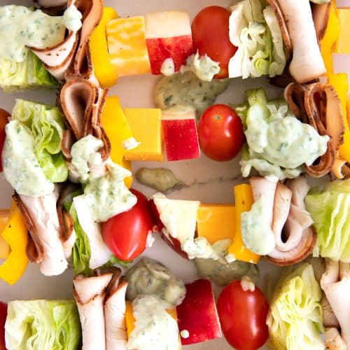 Wooden skewers with repeating layers of cherry tomatoes, iceberg lettuce, sliced Boar's head turkey, cubes of cheese, bell pepper, and cubes of Jazz apples topped with globs of basil mayonnaise.