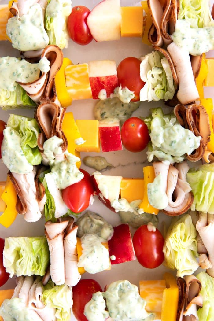 Wooden skewers with repeating layers of cherry tomatoes, iceberg lettuce, sliced Boar's head turkey, cubes of cheese, bell pepper, and cubes of Jazz apples topped with globs of basil mayonnaise.