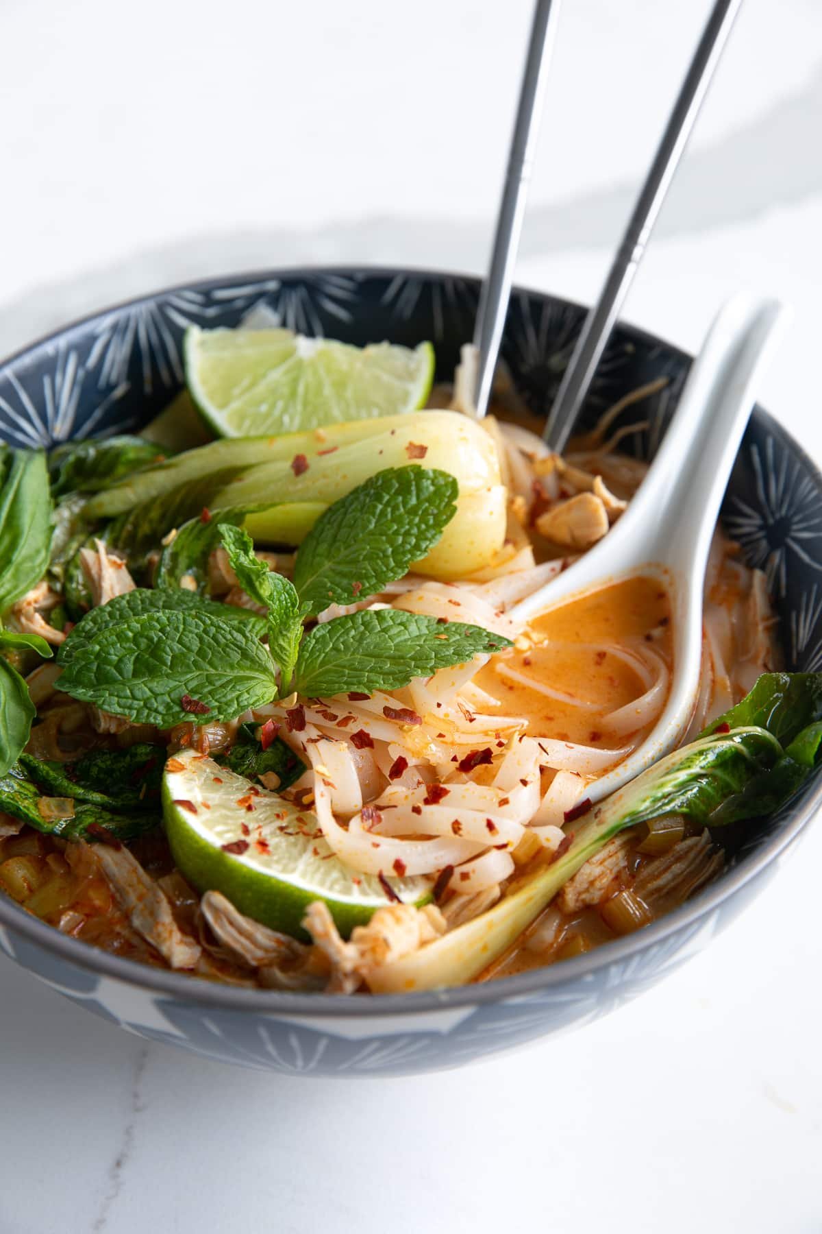 Image of a blue soup bowl filled with Thai chicken curry noodle soup made with a coconut curry broth, rice noodles, shredded chicken, and bok choy, and garnished with fresh mint, limes, and fish sauce.