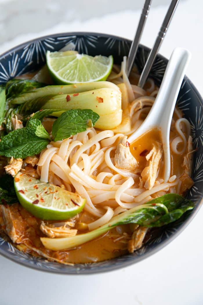 Image of a blue soup bowl filled with Thai chicken curry noodle soup made with a coconut curry broth, rice noodles, shredded chicken, and bok choy, and garnished with fresh mint, limes, and fish sauce.