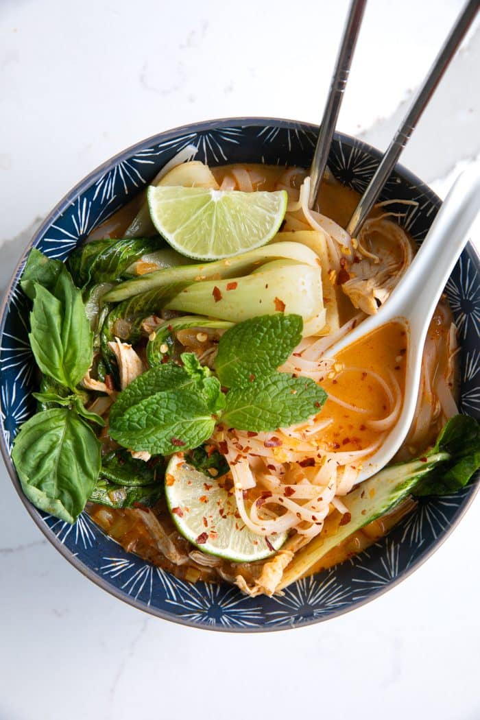 Image of a blue soup bowl filled with Thai chicken curry noodle soup made with a coconut curry broth, rice noodles, shredded chicken, and bok choy, and garnished with fresh mint, limes, and fish sauce.