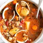 Bowl of Manhattan clam chowder with a tomato-based broth, chunks of potato, carrots, celery, tomatoes, and open clam shells, served with a spoon.