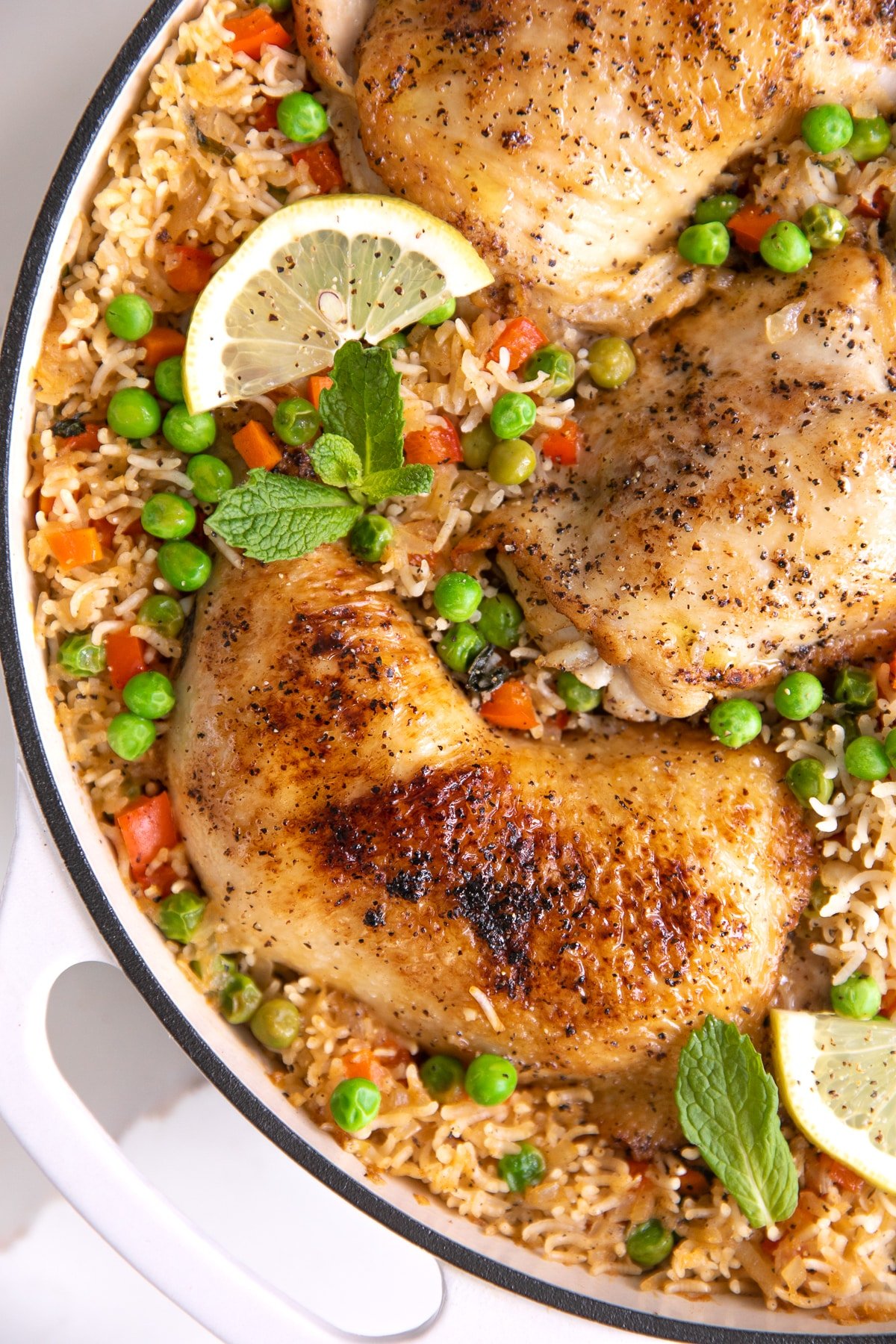 Large white skillet filled with arroz con pollo with peas and finely chopped red bell pepper and garnished with fresh mint and lemon slices.