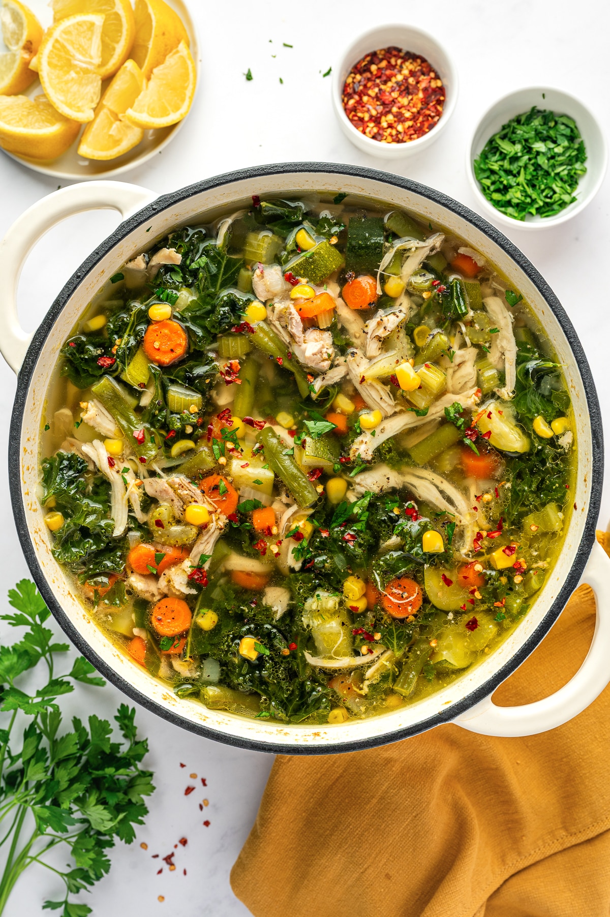 A large white Dutch oven filled with chicken vegetable soup made with shredded chicken, sliced carrots, celery, green beans, corn, zucchini, and kale in a clear golden broth, finished with parsley, black pepper, and red chili flakes.