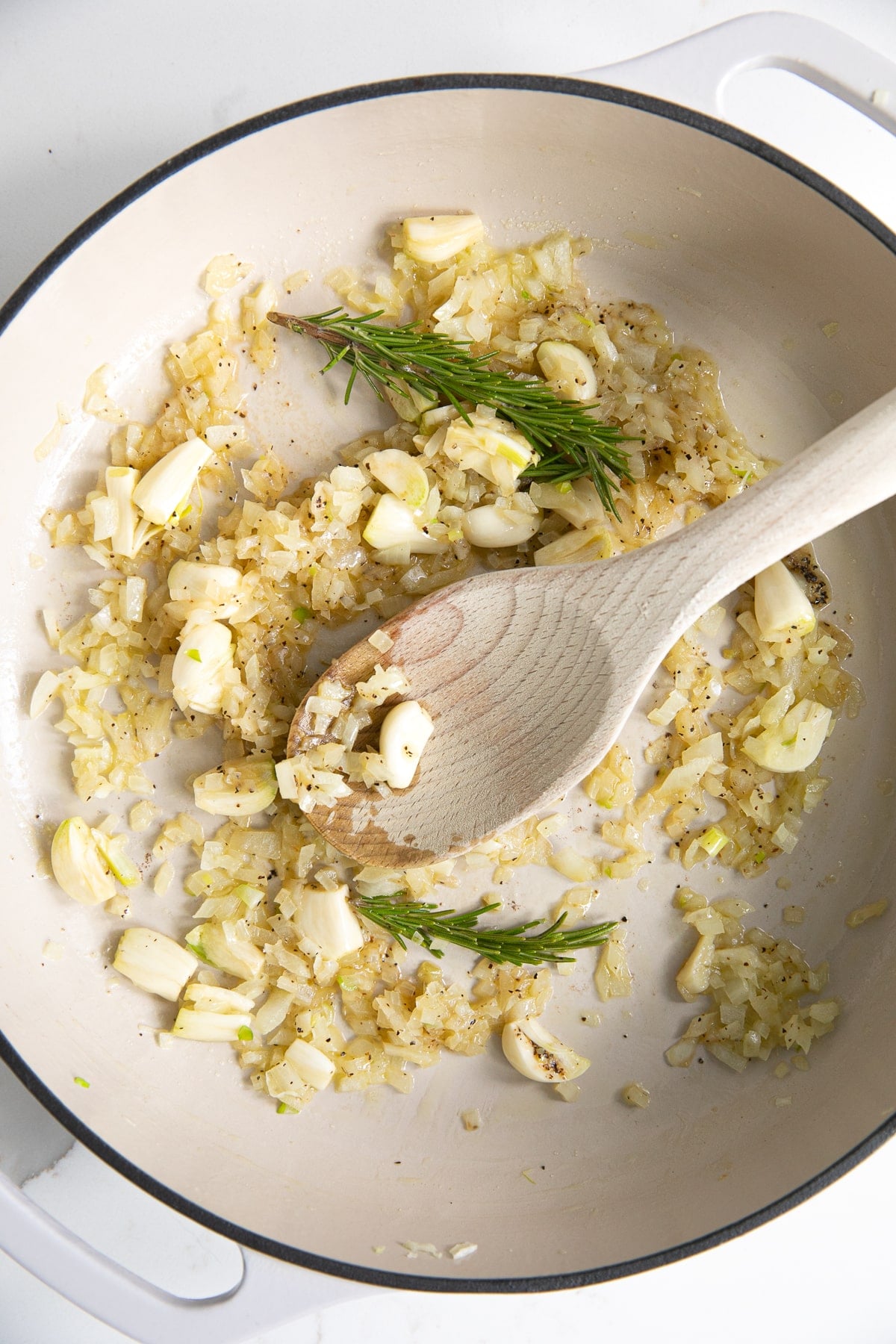 Large white skillet filled with softened diced onion, crushed garlic cloves, and whole sprigs of rosemary.