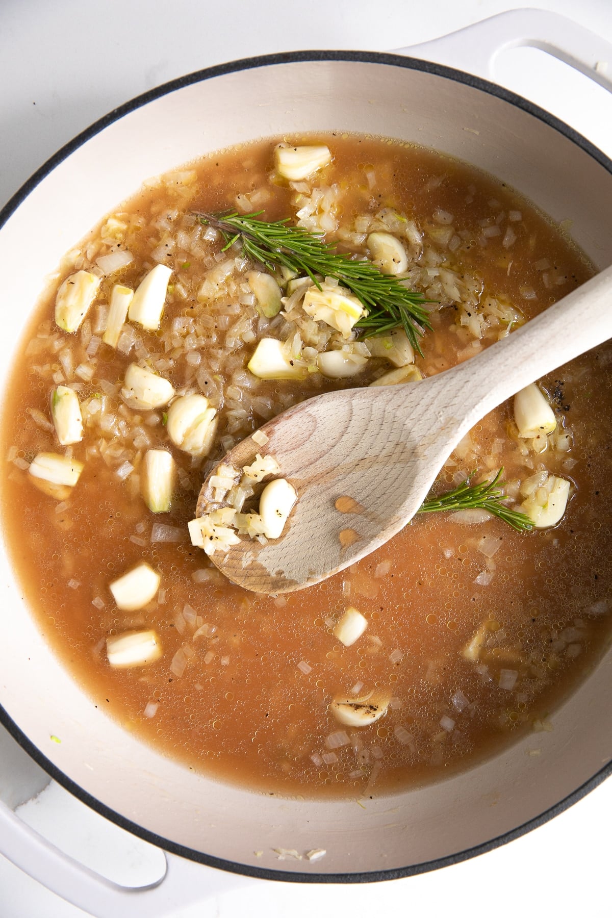 Large white skillet filled with softened diced onion, crushed garlic cloves, and whole sprigs of rosemary cooking in chicken broth and sherry vinegar.
