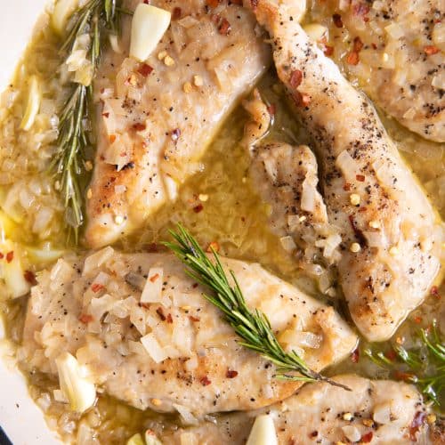 Large white pan filled with thinly sliced chicken breasts simmering in a light garlic and rosemary sauce.