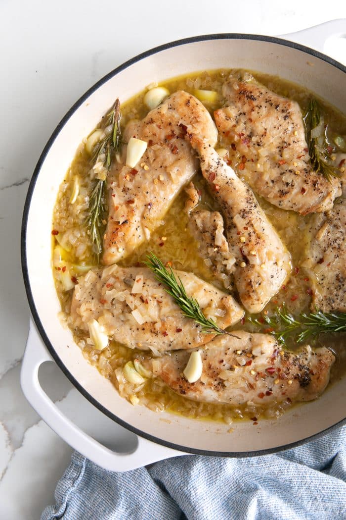 Overhead image of a large white pan filled with thinly sliced chicken breasts simmering in a light garlic and rosemary sauce.