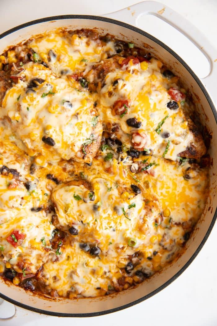 Overhead image of a large white skillet filled with thinly sliced chicken breasts cooked and covered with a mixture of onions, black beans, tomatoes, and sweet corn and smothered in melted cheese.