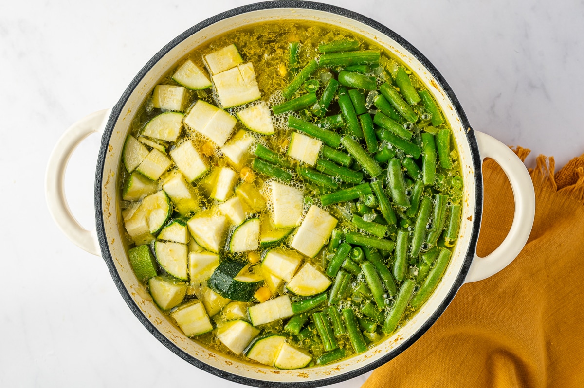 Cut green beans and chunks of zucchini spread across the surface of golden chicken broth in a white Dutch oven after being added later in the cooking process.