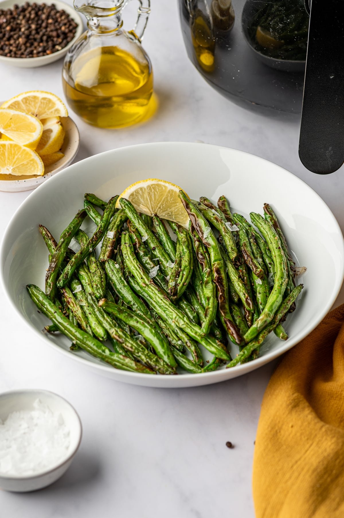Plate of air fryer green beans with lemon slice, olive oil, and peppercorns in the background.