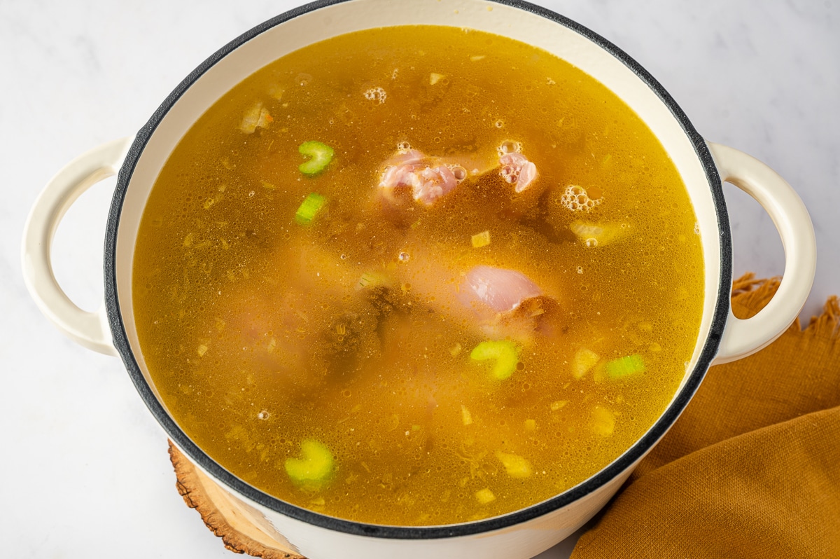 Boneless, skinless chicken thighs submerged in golden chicken broth with onion, celery, garlic, and seasonings in a white Dutch oven as the chicken gently cooks directly in the soup.