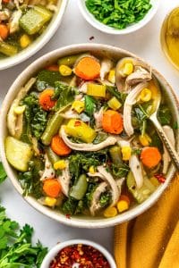 Bowl of chicken vegetable soup filled with shredded chicken, carrots, celery, green beans, corn, zucchini, and kale in golden broth, with a spoon resting along the side of the bowl and fresh parsley and red chili flakes scattered over the soup.