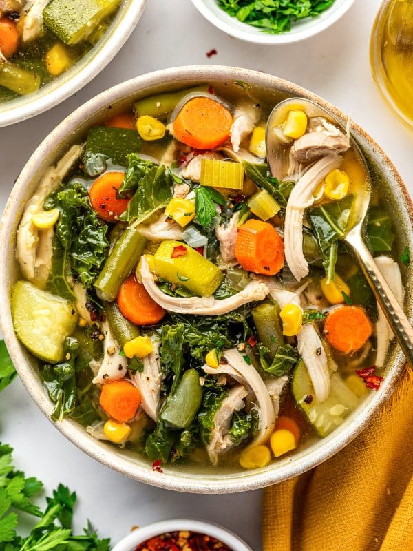 Bowl of chicken vegetable soup filled with shredded chicken, carrots, celery, green beans, corn, zucchini, and kale in golden broth, with a spoon resting along the side of the bowl and fresh parsley and red chili flakes scattered over the soup.