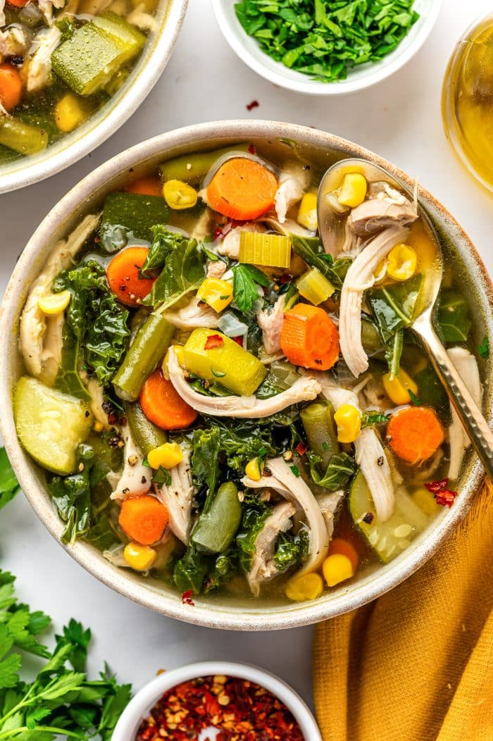 Bowl of chicken vegetable soup filled with shredded chicken, carrots, celery, green beans, corn, zucchini, and kale in golden broth, with a spoon resting along the side of the bowl and fresh parsley and red chili flakes scattered over the soup.