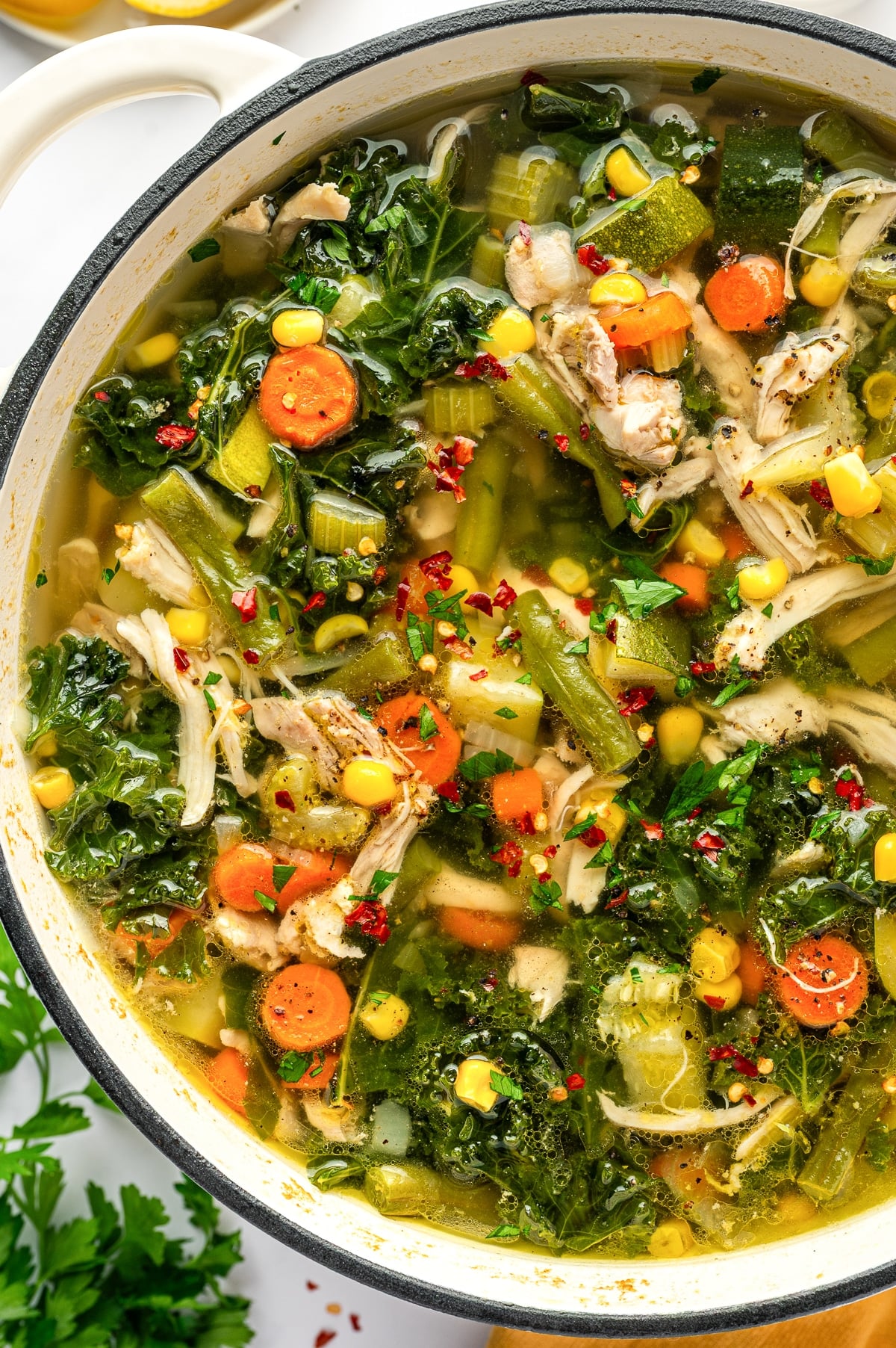 A large white Dutch oven filled with chicken vegetable soup made with shredded chicken, sliced carrots, celery, green beans, corn, zucchini, and kale in a clear golden broth, finished with parsley, black pepper, and red chili flakes.