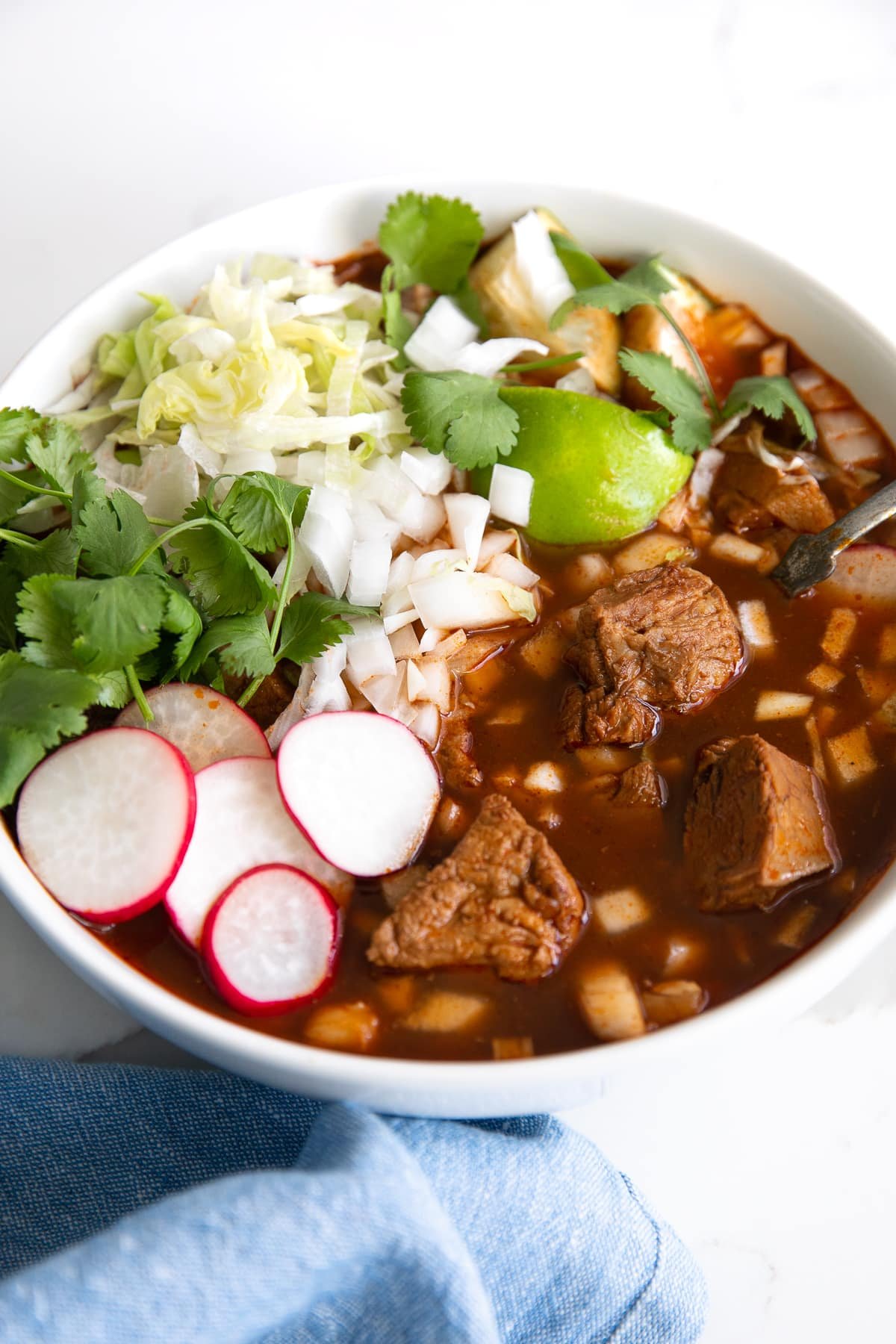 Image of a white bowl filled with posole rojo and topped with cabbage, lettuce, cilantro, and sliced radish.