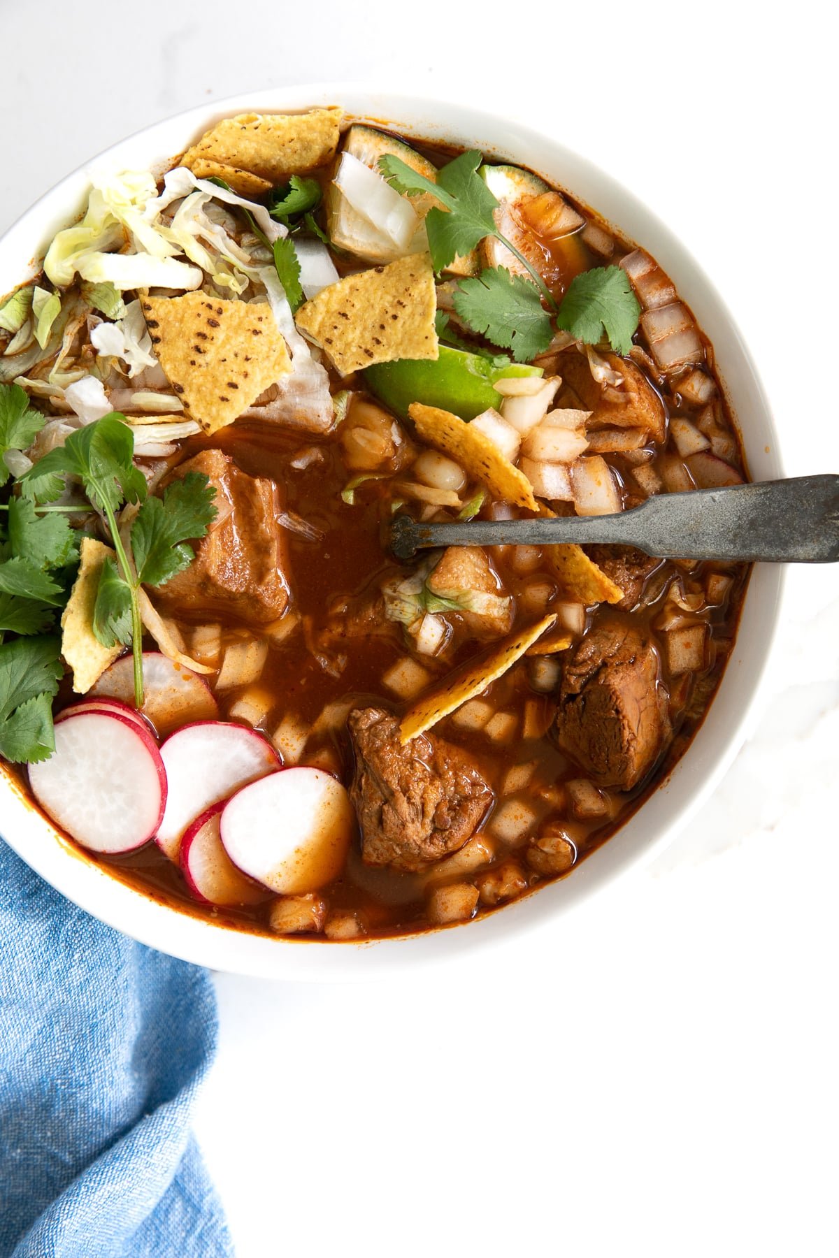 A bowl of posole garnished with shredded lettuce, sliced radish, cilantro, lime, and crushed corn tortillas.