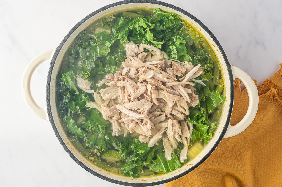A mound of shredded cooked chicken placed in the center of a Dutch oven filled with kale, green beans, zucchini, and chicken broth near the end of cooking.