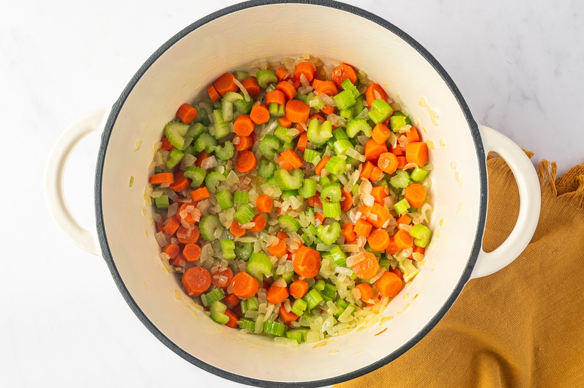 Diced yellow onion, sliced carrots, and chopped celery sauteing together in a white Dutch oven, with the onion beginning to turn translucent and the carrots and celery starting to soften.