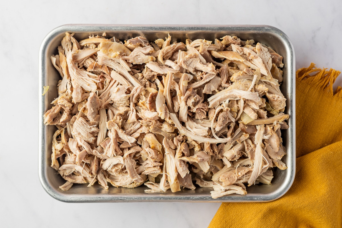 Cooked boneless chicken thighs shredded into irregular bite-size pieces and spread across a rectangular metal tray before being returned to the soup.