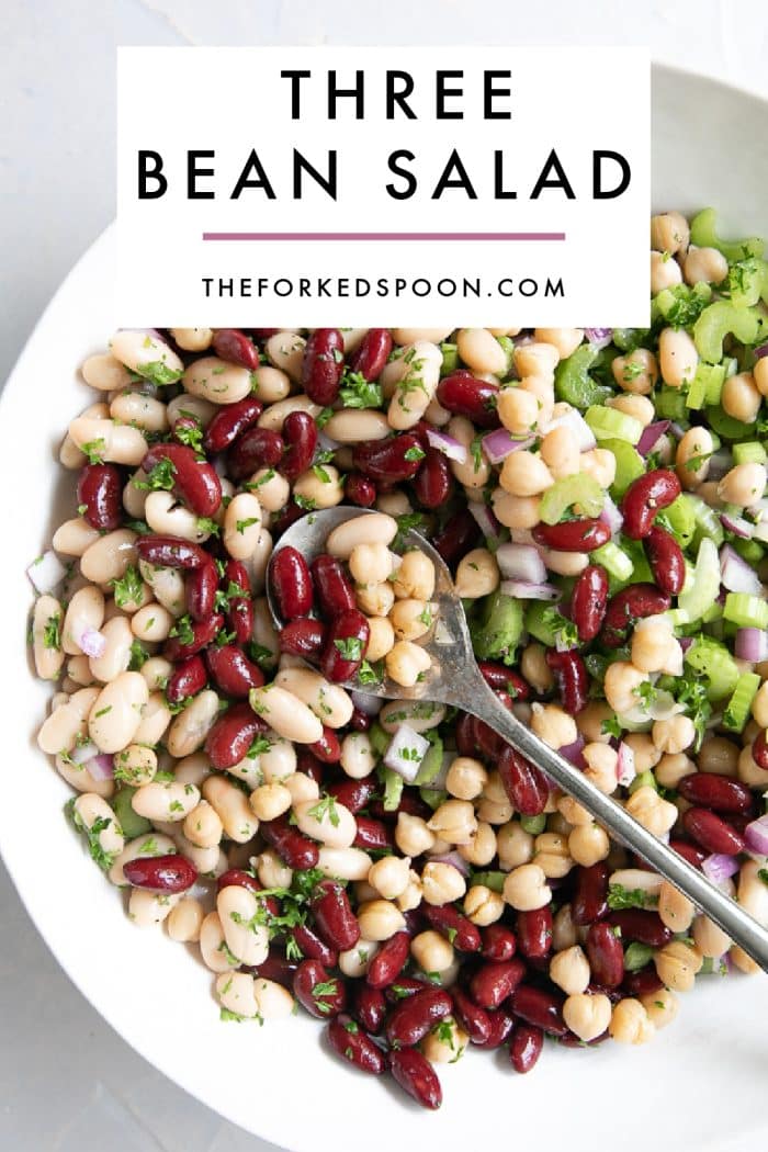 Pinterest pin image for three bean salad showing a white bowl filled with kidney beans, cannellini beans, garbanzo beans, chopped celery, diced red onion, and fresh parsley with a metal serving spoon. A white text box overlay at the top reads โTHREE BEAN SALADโ with โTHEFORKEDSPOON.COMโ underneath and a thin pink line between the title and website.