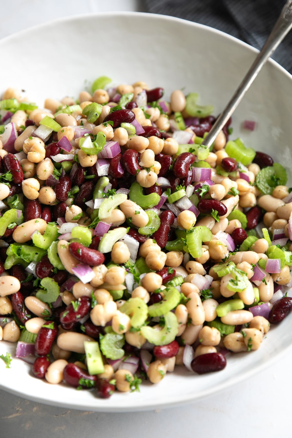 Close-up of three bean salad in a large white bowl with kidney beans, cannellini beans, garbanzo beans, chopped celery, diced red onion, fresh parsley, and black pepper, with a metal serving spoon resting in the salad.