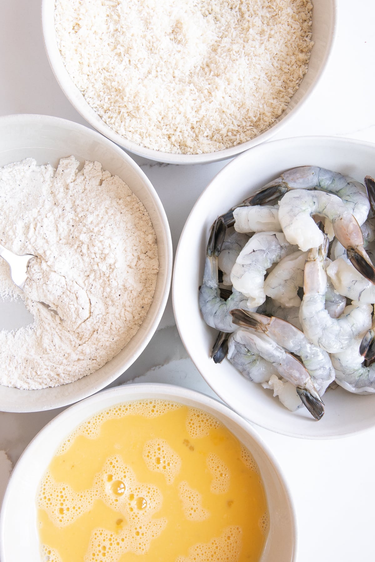 Four white bowls: one filled with raw shrimp, one filled with whisked egg, one filled with flour, and one filled with shredded coconut.