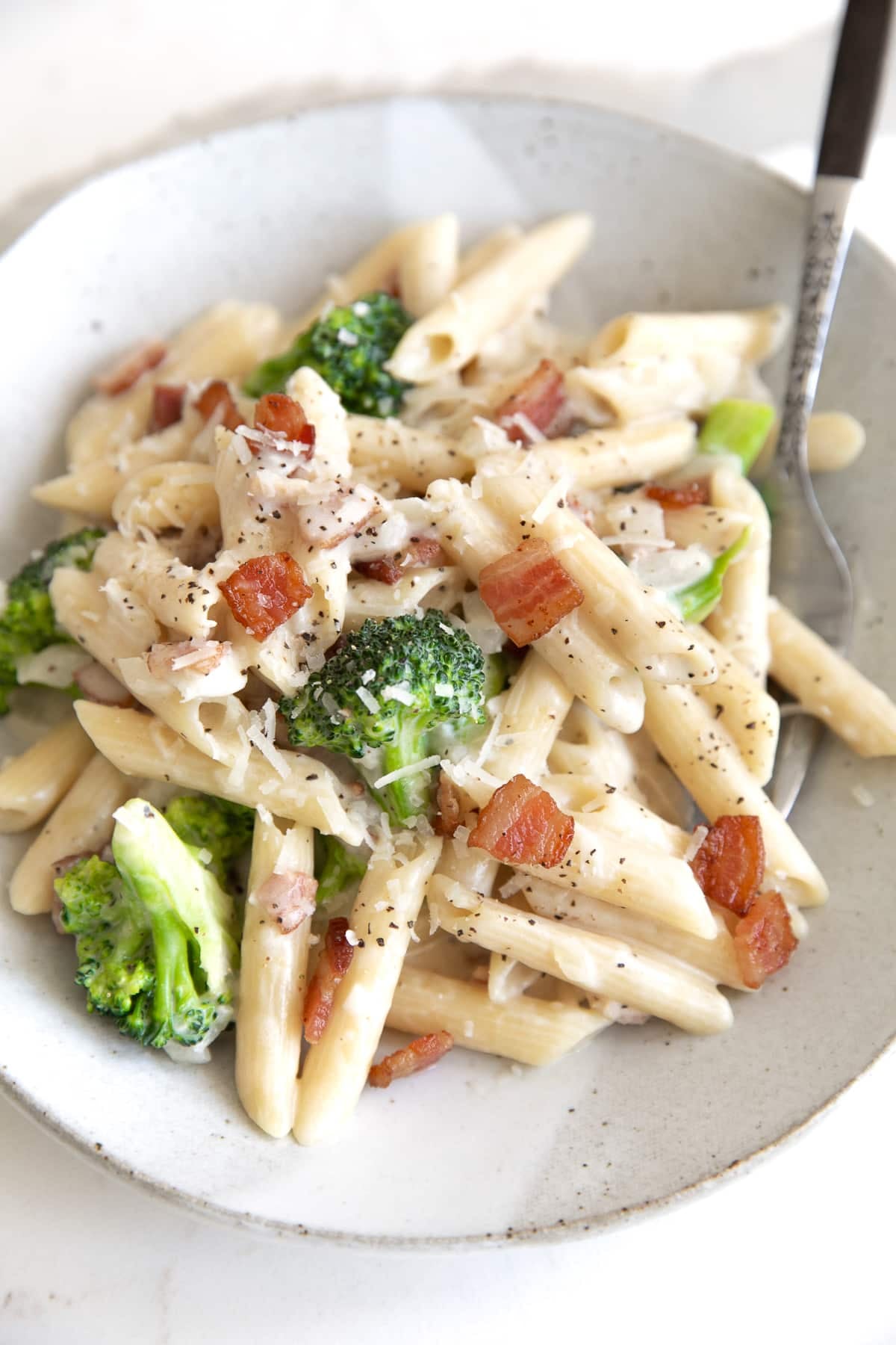 Ceramic serving plate piled with creamy penne pasta filled with small broccoli florets and cooked bacon bits.