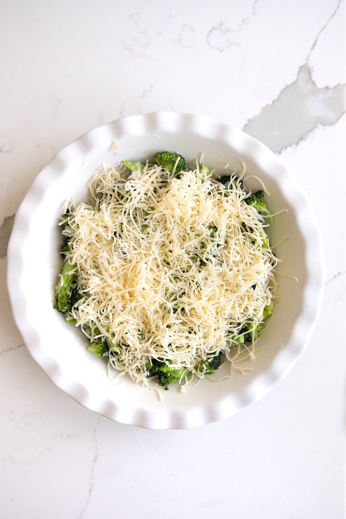 Overhead image of a white pie plate filled with cooked broccoli and shallots and topped with shredded cheese.