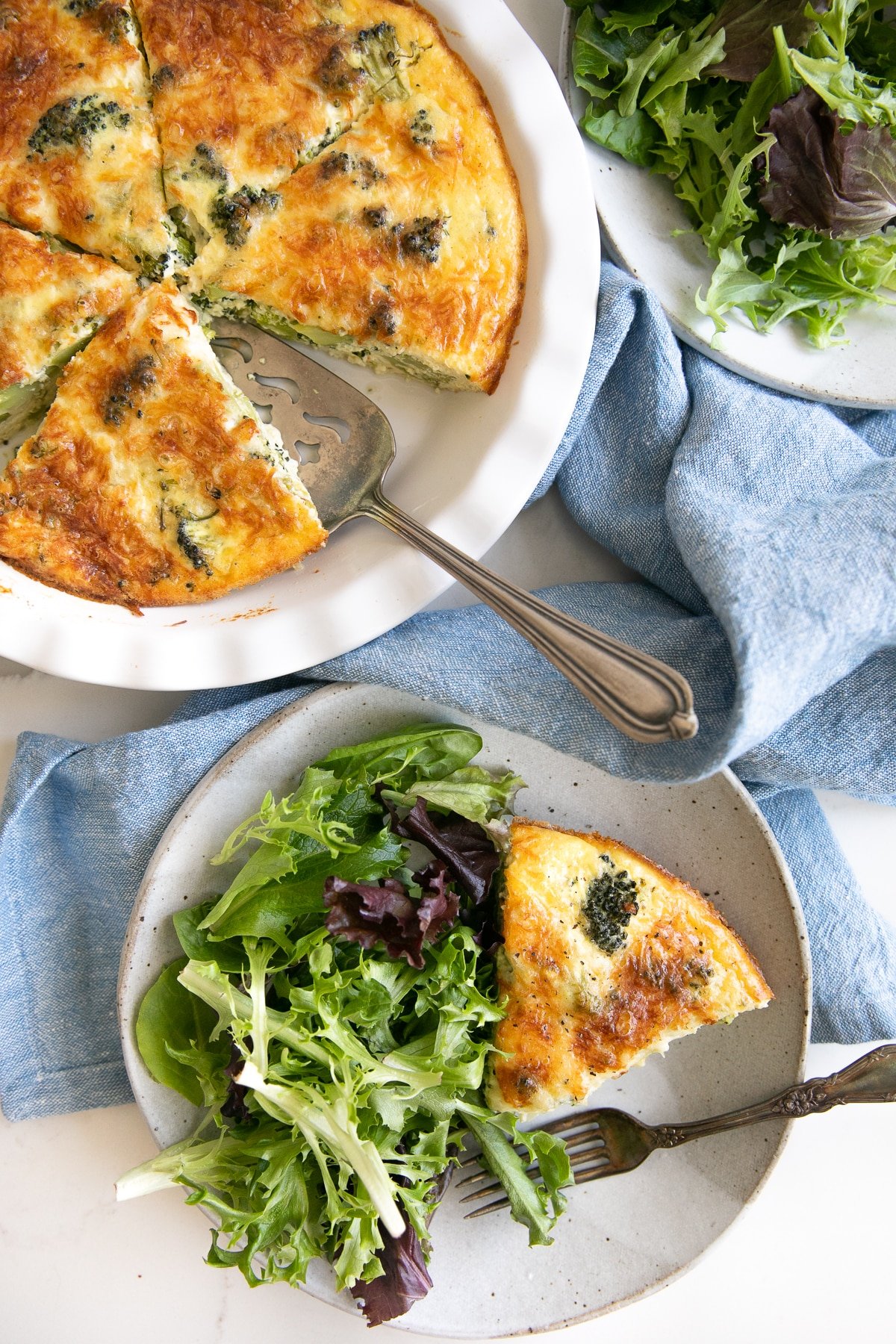 Overhead image of two serving plates filled with a slice of crustless quiche and a side of mixed greens, a blue tea towel, and a white pie dish filled with the remaining quiche.