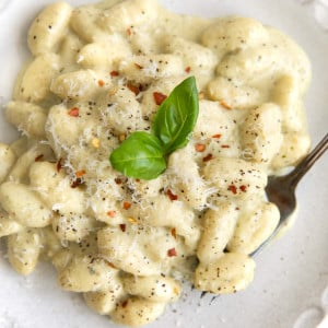 Overhead image of a white serving plate filled with creamy pesto gnocchi garnished with freshly grated parmesan cheese, red chili flakes, and fresh basil leaves.
