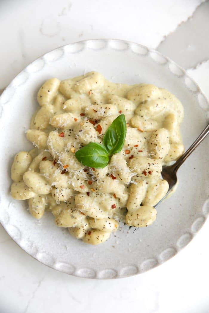 Overhead image of a white serving plate filled with creamy pesto gnocchi garnished with freshly grated parmesan cheese, red chili flakes, and fresh basil leaves.