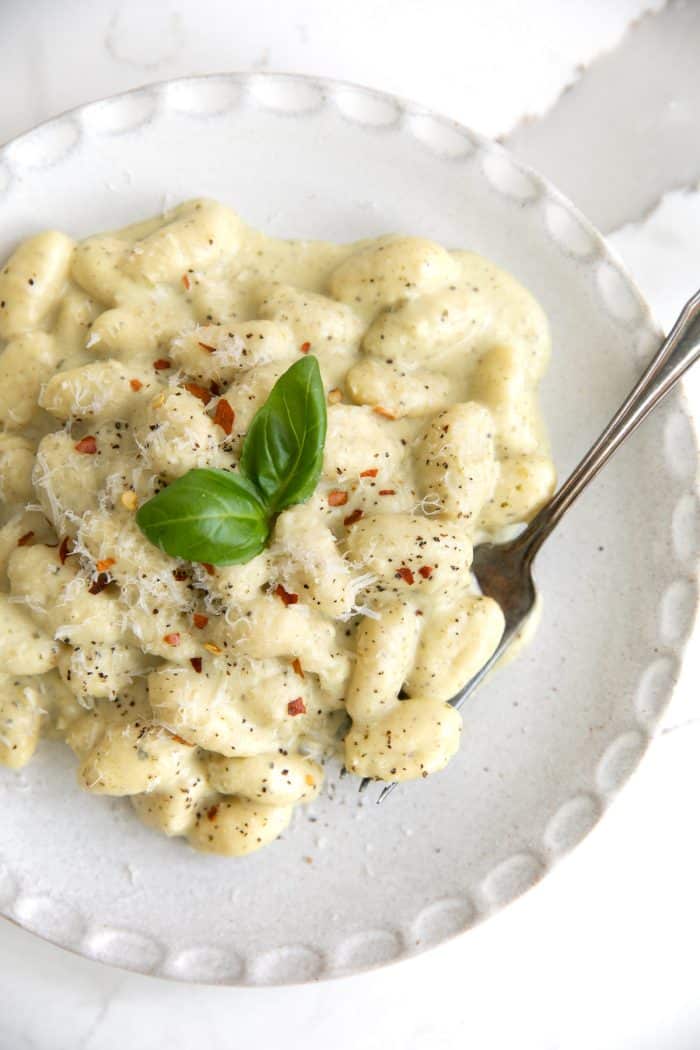 Image of a white serving plate filled with creamy pesto gnocchi garnished with freshly grated parmesan cheese, red chili flakes, and fresh basil leaves.