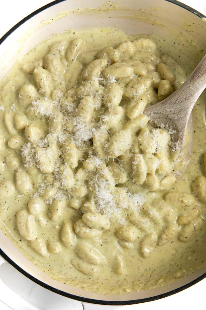Overhead image of a white ceramic pan filled with cooked gnocchi in a pesto creamy sauce and covered with freshly grated parmesan cheese.