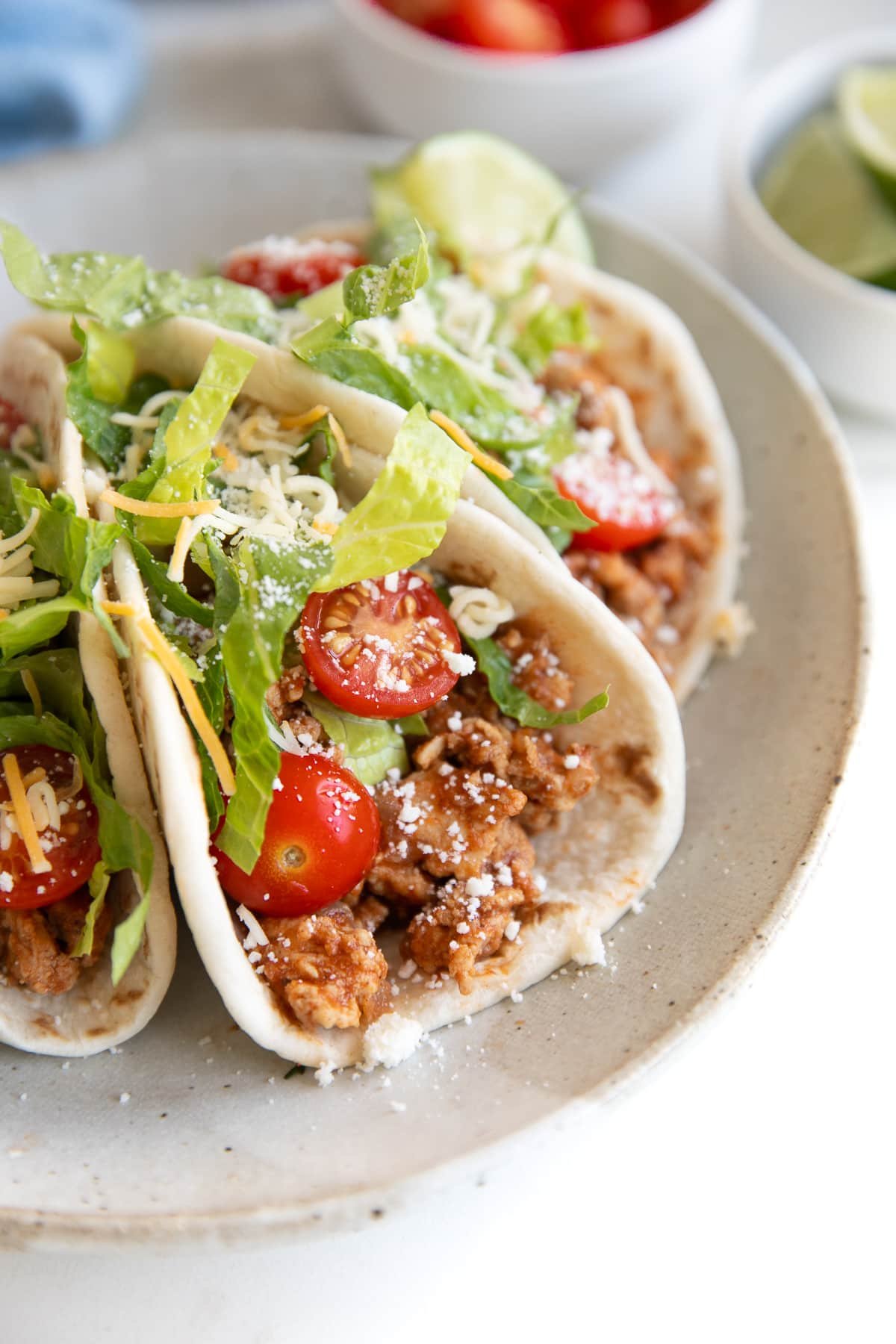 Three ground turkey tacos filled with ground turkey taco meat and topped with tomatoes, lettuce, and cheese, on a small serving plate.