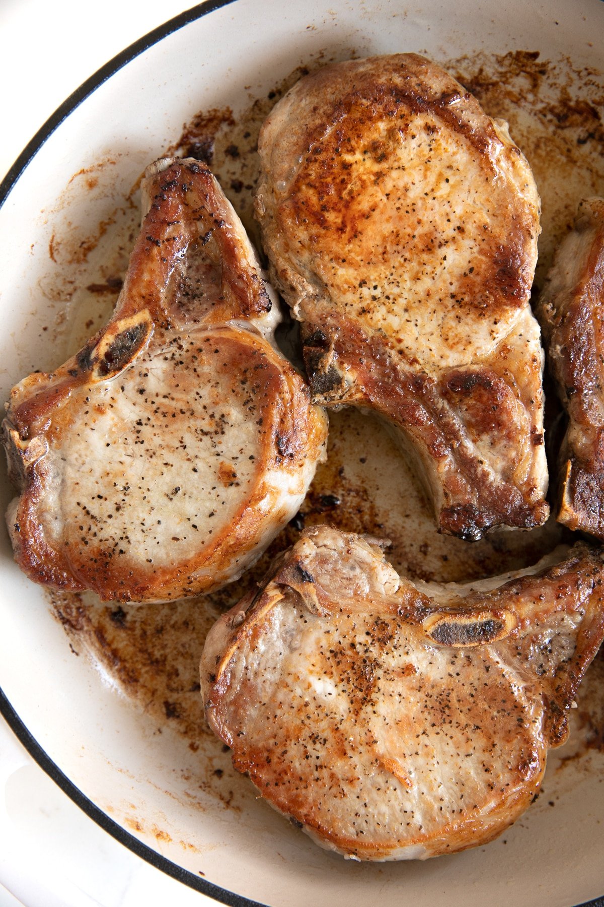 Large white skillet filled with four bone-in pork chops seared in butter.