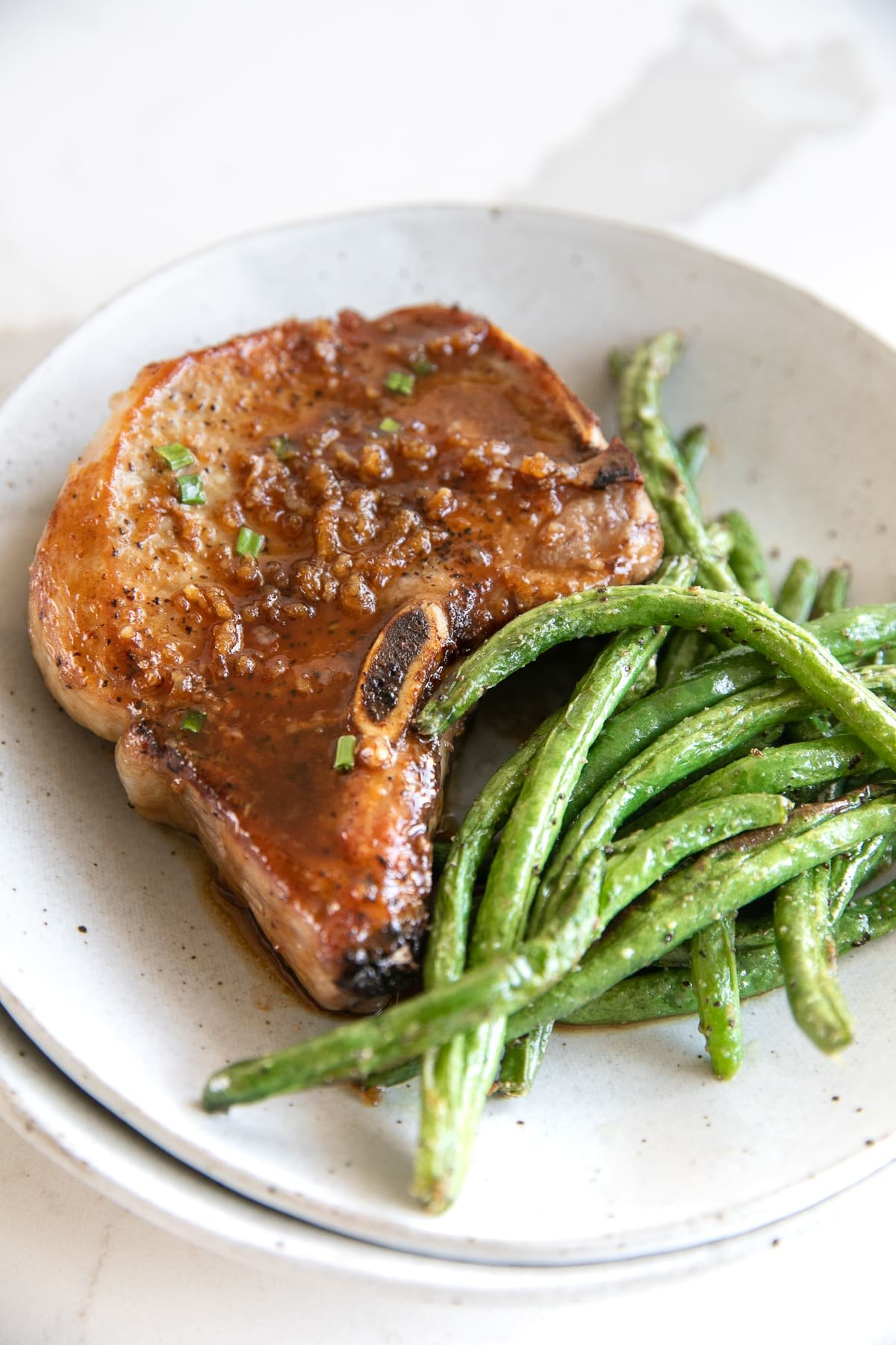 White serving plate filled with one bone-in honey garlic pork chop and a side of green beans.